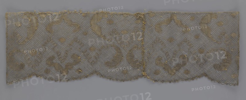 Lace fragment, lace, 18th century, Europe, fabric, width 20 cm, length 120 cm, A rectangular piece of delicate openwork fabric with a net-like ground and an embroidered scrolling pattern, shown against a plain background. The textile displays a scalloped lower edge and a straight upper edge, with an apparent seam or join near the center. The embroidery and net appear in muted tones of gold and beige, while the background surface beneath the textile shows a cool gray. The fabric has areas of slight fraying and a few loose threads along the edges and seam. The overall visual effect is a repeating ornamental design of curved paisley-like motifs and small round fills across the netted field.