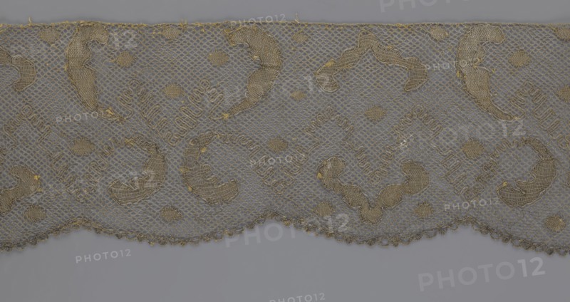 Lace fragment, lace, 18th century, Europe, lace, fabric, width 20 cm, length 120 cm, A strip of delicate lace in shades of gray and gold fills the upper portion of the image, featuring a repeating scalloped edge and a net-like background; the lace has several ornate, curved embroidered motifs and small circular adornments in metallic gold thread, with tiny loose threads and slight fraying visible along some edges; the lower portion shows a smooth gray surface beneath the lace.