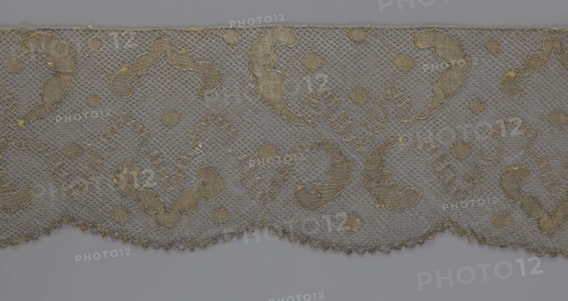 Lace fragment, lace, 18th century, Europe, lace fabric, width 20 cm, length 120 cm, A piece of lace fabric with a net-like background and a scalloped lower edge, featuring a repeating pattern of curved and diamond-shaped embroidered motifs in muted gold and tan threads against a soft gray background, with a few small embroidered dots and subtle variations in thread thickness and texture.