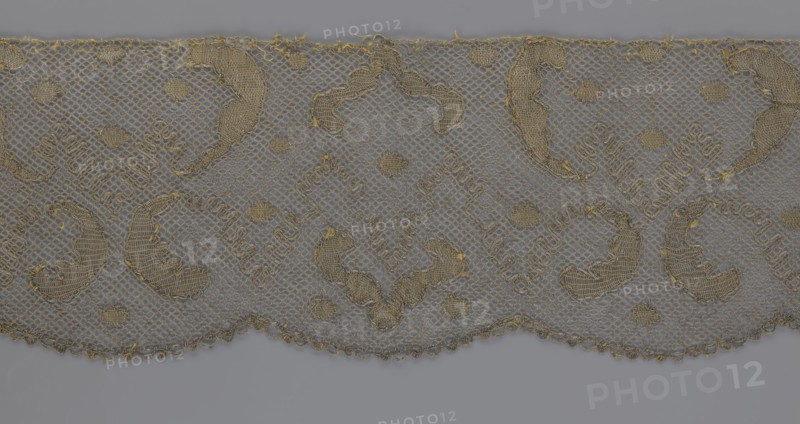 Lace fragment, lace, 18th century, Europe, fabric, width 20 cm, length 120 cm, A piece of lace fabric with a repeating decorative pattern, showing a scalloped lower edge and a straight upper edge, woven in a mesh net structure with embroidered motifs; the lace appears in shades of beige and gold against a gray background, with areas of denser stitching forming curved and circular shapes, small rounded fillers, and ornamental loops, and with visible thread details and fringe along the top edge.
