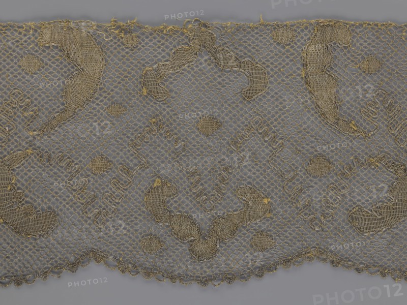 Lace fragment, lace, 18th century, Europe, lace fabric, width 20 cm, length 120 cm, A section of lace fabric with a netted background in gray and metallic gold threads forming an all-over pattern, showing a scalloped lower edge and a straight upper edge, with several larger solid embroidered motifs and a few smaller rounded filled motifs distributed across the surface, the embroidery creating textured raised areas and loose thread ends in places, overall colors visible are gray and gold.
