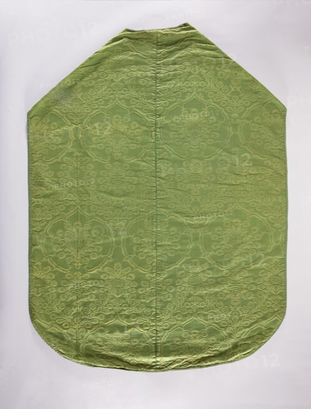 Chasuble;, 16th century, Italy. Liturgical vestment church paraments, length 129 cm, width 97 cm back. Design: plant ornaments and flowers — a seven-leafed rose with a floral thistle flower; above the rose is a small crown and above that the same floral thistle motif. From the crown two intertwined floral branches branch down, creating a roof-like form; this motif repeats in horizontal rows. Trimmed with a narrow green ribbon; green cotton lining. Function: sacred art, liturgical vestment for churches and chapels. Repossession, A single large fabric panel laid flat with a pointed top and rounded bottom, showing a subtle raised floral and scroll damask pattern across its surface; the fabric appears primarily light green with slightly darker green raised motifs and gentle wrinkles and seams running vertically; the background behind the panel is a pale off-white.