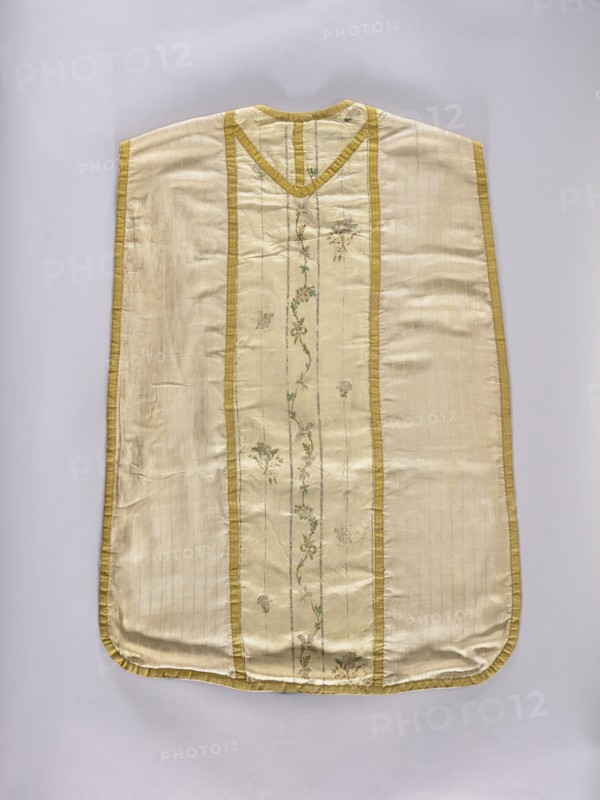 Chasuble, Liturgical vestment made of three types of silk fabrics, trimmed with yellow silk braid; cotton lining green on the front, navy blue on the back. Front features an embroidered column of white silk grosgrain embroidered with silk threads in patterns of small floral sprigs; three sprigs placed sparsely, likely cut from another piece and added. Back column and neckline made of silk weft-weave fabric brocaded with silk yarn and silver thread in a striped pattern with a wavy floral tendril and small floral sprigs. Sides made of cream-colored striped silk fabric. Decorated with flowers, leaves, and plant ornaments. 2nd half of the 18th century. Place of origin: Poland; back: France. Length 98.5 cm, width 68 cm., A flat garment displayed against a pale background, made of a shiny cream fabric with a central vertical panel bordered by narrow golden-yellow trim and decorated with a delicate vertical vine of embroidered small flowers and leaves in muted green and pale brown tones; the ...