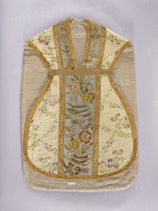 Chasuble, Chasuble made of cream-colored silk brocaded with a pattern of leafy, twisting twigs bearing small fruits and rose blossoms; brocade threads in pink, blue, and green. Front and back columns embroidered with cornucopia motifs producing bouquets of flowers, including tulips and roses; gold background of the columns, ornament in pink, yellow, and blue. Gray linen lining. Mid-18th century, Poland. Liturgical vestment. Length 112 cm, width 72 cm back., A textile garment with a symmetrical, sleeveless shape is shown flat against a pale background; the main fabric is a light beige or tan with panels of cream or ivory silk featuring embroidered floral motifs, and a central vertical panel with multicolored woven flowers and leaves. Several narrow bands of gold trim run vertically and outline the panels, creating a framed pattern, while the embroidered flowers include shades of pink, green, blue, brown, and yellow. The overall palette includes beige, cream, gold, pink, green, blue, yel...