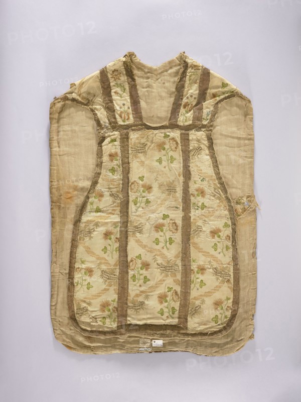 Chasuble, embroidered on white rep with pastel-colored silks and gold thread in patterns imitating brocade: wavy ribbons and floral bouquets forming a network arrangement; columns emphasized with gold galloon stitching; cotton lining; embroidery colors: green, ochre, beige, blue. Liturgical vestment, 2nd half of the 18th century, Poland. Length 96 cm, width 66 cm back., Keywords: liturgical vestments, flowers, plant ornaments, church paraments, sacred art, churches and chapels furnishings., A sleeveless garment laid flat against a pale background, primarily in shades of beige and cream with a patterned central panel featuring floral motifs in green and muted brownish-pink tones; the garment has vertical and curved bands in a darker tan or brown framing the patterned sections, and visible frayed edges and worn areas with some discoloration in orange-brown; the fabric appears textured with a slightly shiny finish in places and a matte backing in others, showing small irregularities and r...