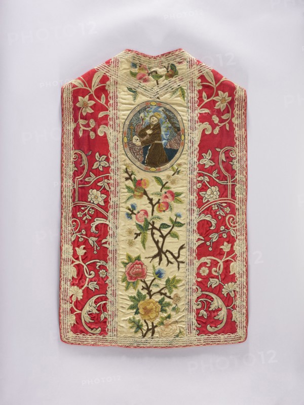 Chasuble, 17th century, Poland. Liturgical vestment, length 95 cm, width 60 cm back. Cotton lining. Trimmed with period white lace. Predominant colors: white for the columns, red for the sides, and yellow, pink, blue, and white for the ornament. Decoration: columns with plant ornaments — at the back, at the top in an oval embroidered kneeling St. Anthony Anthony of Padua, 1195–1231, below branches of apple blossoms and fruit; at the front, branches with apple blossoms and fruit. Sides: ornament with stylized plant twigs. Iconography: Anthony of Padua; function: church paraments / sacred liturgical vestment., An ornate textile panel with a central cream-colored vertical band framed by a decorative lace trim, flanked by wide red panels patterned with pale floral and scroll embroidery; the cream band features embroidered flowering branches with pink, yellow, blue, and green floral motifs and a circular embroidered medallion near the top showing a figure in brown robes against a blue and t...