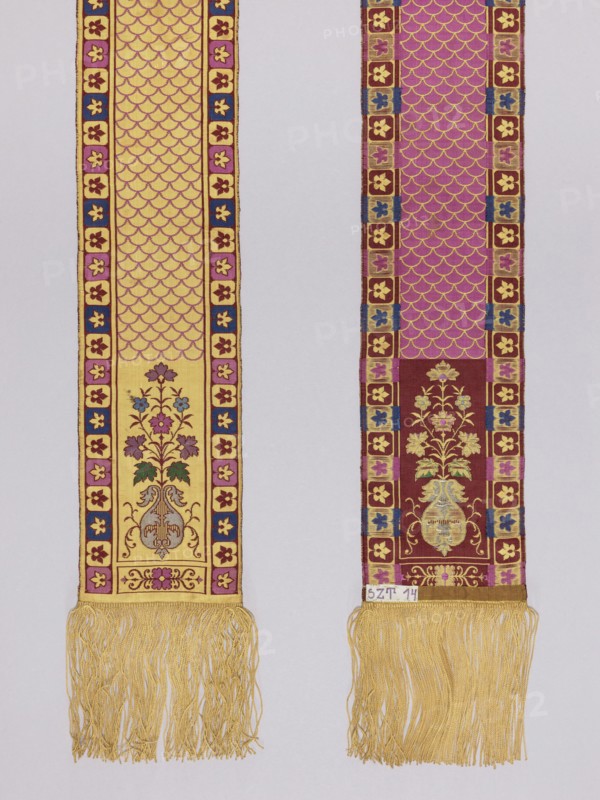 Half-belt. Double-sided silk half-belt. Each head features a single motif of a flower bouquet in a vase. Center with a carp scale motif. Vertical borders divided into small quarters, each with leaf motifs; the lower horizontal borders upper ones missing contain single rosettes. Borders in the form of straight lines. Background colors: yellow and brown; ornament colors: blue, purple, yellow, green, silver. Fringe sewn on; materials: silk, straw. Weave density approx. 18/25 x2. Date: 1890–1910. Function: clothing accessory, national costume; length 345 cm; width 12 cm. Polish kontusz/double-sided belt style; men's clothing accessory. Plant ornaments, rosette, carp scale ornament., Two long vertical woven bands hang side by side against a pale background, each ending in a fringe of golden yellow threads. The left band features a golden yellow field with an allover scalloped pattern in a darker tone and a narrow border of small floral motifs in pink, blue, and maroon; near its lower sectio...