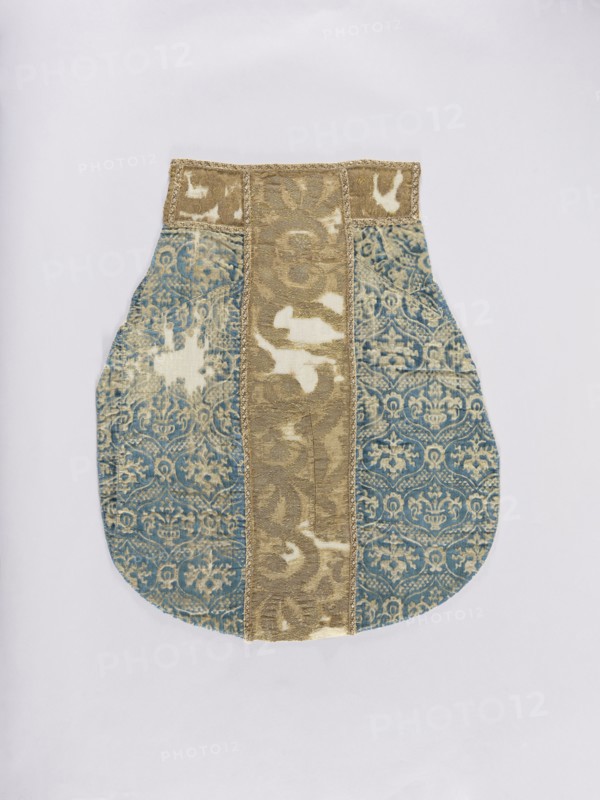 Chasuble, liturgical vestment decorated with flowers, leaves, and plant ornaments; church paraments, sacred art. Chasuble of Genoese velvet, patterned, convex, with a regularly repeating motif of a stylized bouquet of flowers in vases enclosed in a thick heart-shaped net; mesh fastened with palmettes. Colors: sapphire blue ornament on cream-gold background; columns of solid material in gold-beige. Ornamentation on fabric is not fully legible due to large defects. Made of stylized plant forms. 15th/16th century; possibly Italy or France, sides: Genoa. Length 122 cm, width 64 cm back., An object resembling a shaped textile panel lies flat against a pale background; it has a rounded lower body with a narrower rectangular top and is made from patterned fabric with a central vertical strip in a muted gold color flanked by two wider blue panels featuring an ornate repeating floral and scroll motif, with areas of lighter worn patches on both the gold and blue fabrics; the top edge includes a ...
