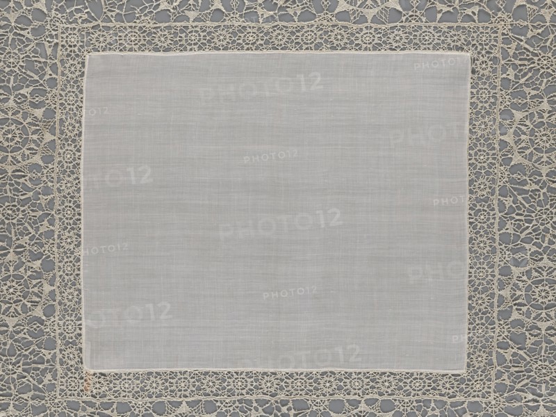 Doily, geometric ornaments; rosettes ornament; doilies. Smooth center. Edge made of white linen lace with rosette motifs: large rosettes in the center and rows of small rosettes on the edges; punto in tela lace; ca. 1600, Italy. Materials: lace, fabric. Height 79 cm, width 70 cm., A square piece of light gray fabric with a smooth, slightly textured surface sits centered within an ornate openwork border; the border is a delicate lace in an off-white or cream color featuring repeating circular and web-like motifs, and it contrasts with a darker gray background visible through the lace openings.