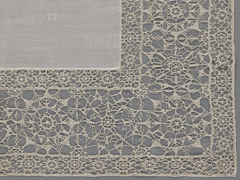 Doily, geometric ornaments, rosettes ornament, doilies. Smooth center. Edge made of white linen lace with rosette motifs: large rosettes in the center and rows of small rosettes on the edges, punto in tela lace, ca. 1600, Italy. Lace, fabric; height 79 cm, width 70 cm., The image shows a section of lace fabric with a solid woven square of cloth in the upper left corner bordered by an intricate openwork lace design. The lace is composed of a network of delicate loops and starburst-like motifs, forming geometric and floral patterns connected by fine threads. The visible colors are shades of off-white or cream for the fabric and lace, contrasted against a muted gray background that shows through the open spaces. The textures range from the smooth, tightly woven cloth to the raised, airy, and filigree-like texture of the lace.