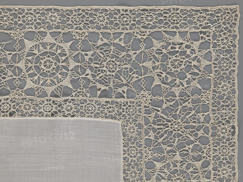 Doily, geometric ornaments; rosettes ornament; doilies; smooth center. Edge made of white linen lace with rosette motifs: large rosettes in the center and rows of small rosettes on the edges; punto in tela lace; ca. 1600, Italy; lace fabric; height 79 cm, width 70 cm., A close-up view of a corner of a rectangular textile featuring a solid woven central panel in a pale off-white color bordered by an intricate openwork lace in a slightly warmer creamy off-white; the lace displays repeating geometric and circular motifs connected by delicate looping threads, with visible small open spaces throughout, and the background beneath the lace appears as a muted medium gray.