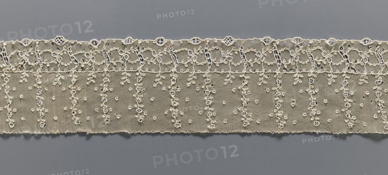 Fragment of lace, lace, ornament: continuous vertical twigs at the top with points between them; at the bottom a strip of edge ornament where plant and geometric tendrils intertwine. Late 18th century, possibly Germany or France. Fabric lace, width 9.5 cm, length 67 cm., A horizontal strip of lace in an off-white or cream color stretches across a gray background, featuring a scalloped top edge with a row of small circular openings and an ornate looping embroidered border beneath; below that are several vertical braided or beaded-looking embroidered columns ending in tiny leaf-like motifs, set against a fine netted ground sprinkled with a few small raised circular dots and delicate embroidered clusters, with the lower edge of the lace slightly scalloped and uneven.