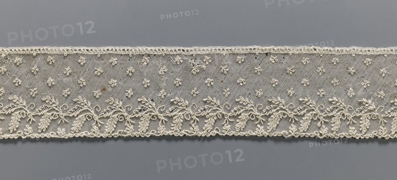 Fragment of lace, Alençon lace, decorated with small twigs in projections and a tendril with feathery leaves running parallel to the edge against a mesh background, 2nd half of the 18th century, France, lace, width 8.5 cm, length 54 cm, A horizontal strip of delicate lace in an off-white or cream color stretches across a neutral gray background; the lace features a fine hexagonal mesh filled with a scattered pattern of small rosette-like motifs and a continuous embroidered border along the lower edge composed of sinuous vine shapes with leafy clusters and curled tendrils, while the top edge has a narrow scalloped trim; the overall texture appears intricate and slightly raised against the smooth gray surface.