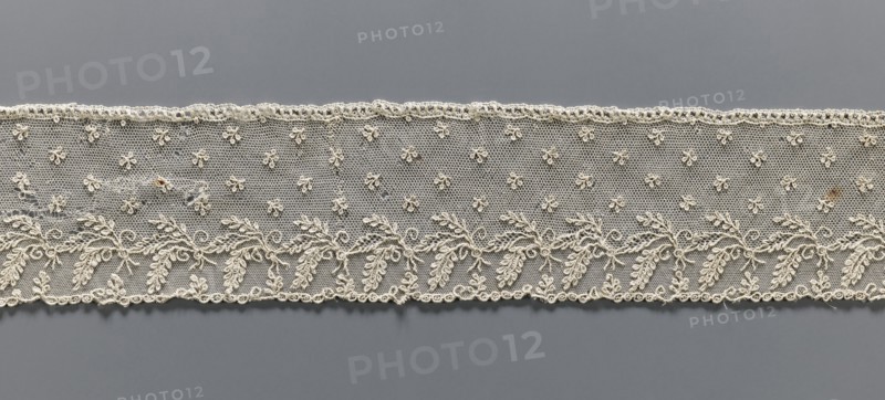 Fragment of lace, Alençon lace, decorated with small twigs in projections and a tendril with feathery leaves running parallel to the edge against a mesh background. 2nd half of the 18th century, France, lace, width 8.5 cm, length 58.5 cm., A horizontal strip of delicate lace in off-white and cream tones stretches across a gray background, featuring a fine netted field with a pattern of small floral motifs and a lower border of intertwined leafy garlands and scrolls, with scalloped edges and a few small areas of darker discoloration.