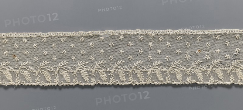 Fragment of lace, Alençon lace decorated with small twigs in projections and a tendril with feathery leaves running parallel to the edge against a mesh background; 2nd half of the 18th century, France, lace; width 8.5 cm, length 58.5 cm., A horizontal strip of delicate lace in off-white spans the image against a flat gray background, with a pattern of small embroidered floral motifs on a fine hexagonal mesh above a band of more densely stitched leafy vine designs; the lace edge shows a scalloped trim and a few loose threads, and there are a few small darker brown spots on the fabric.