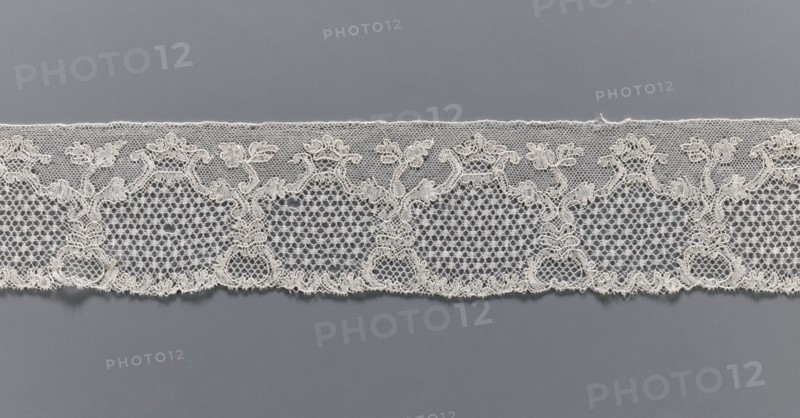 Lace fragment, purchased provenance noted, lace, Malines Mechelen lace, shell and plant pattern; mesh background: point de neige and drochelle; 2nd half of the 18th century; Mechelen; lace; width 7.7 cm, length 521 cm, A horizontal strip of delicate lace laid against a smooth background, showing an intricate repeating floral and scrollwork pattern with a scalloped lower edge and a straight upper edge; the lace has open hexagonal mesh sections interspersed with denser embroidered motifs that form stylized flowers and curving vines, and the piece appears in shades of off-white and cream against a medium gray backdrop.