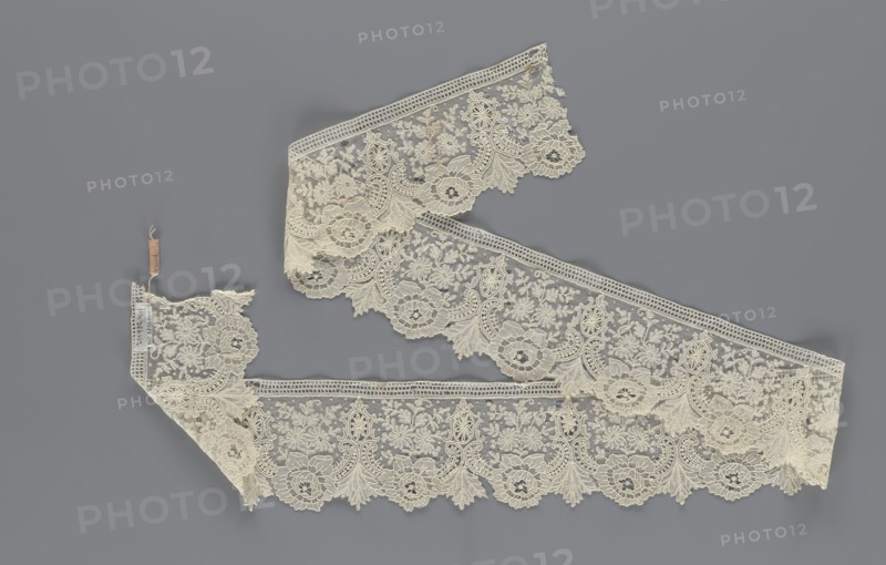 Lace fragment, lace, plant ornaments. Serrated on one side, with a pattern of flowers, fan-shaped leaves, and ovoid rosettes above them. Rose flowers between the leaves, and sprigs of other small flowers above them. Background fond de gaze. 2nd half of the 19th century, Brussels, lace, width 8.5 cm, length 127 cm, A length of delicate lace trim displayed against a gray background, the lace is an off-white or cream color with intricate floral and ornamental patterns, scalloped edges, and a fine mesh ground; several sections are arranged in a loose zigzag, with a few small attached tags visible at one end; the design shows repeating round floral motifs, leafy sprays, and ornamental scrolls, with textured raised embroidery and openwork areas.