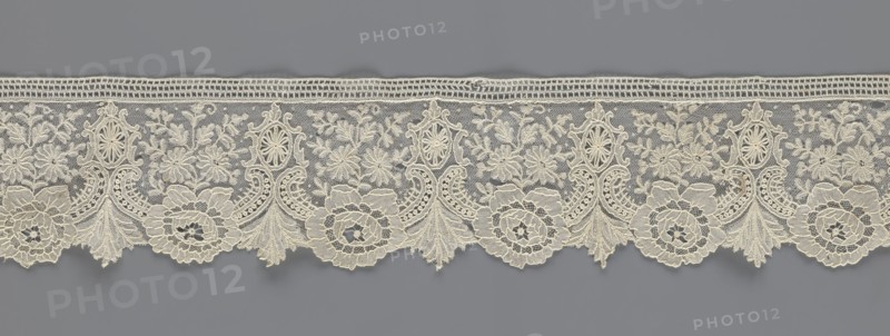 Lace fragment, lace with plant ornaments. Serrated on one side, with a pattern of flowers, fan-shaped leaves, and ovoid rosettes above them. Rose flowers between the leaves, and sprigs of other small flowers above them. Background fond de gaze. 2nd half of the 19th century, Brussels; lace; width 8.5 cm, length 127 cm., A horizontal band of delicate lacework with an ornate repeating floral and scroll pattern, displayed against a muted gray background. The lace is a pale cream color and features a scalloped lower edge with a few large circular floral motifs, several fan-shaped leaf elements, and a group of smaller flowers and leaves woven into a fine net ground. The upper edge has a narrow openwork border with a rectangular mesh. The overall texture appears intricate and raised in places, showing loops, cords, and denser embroidered areas contrasting with the sheer net. The visible colors are cream and gray.