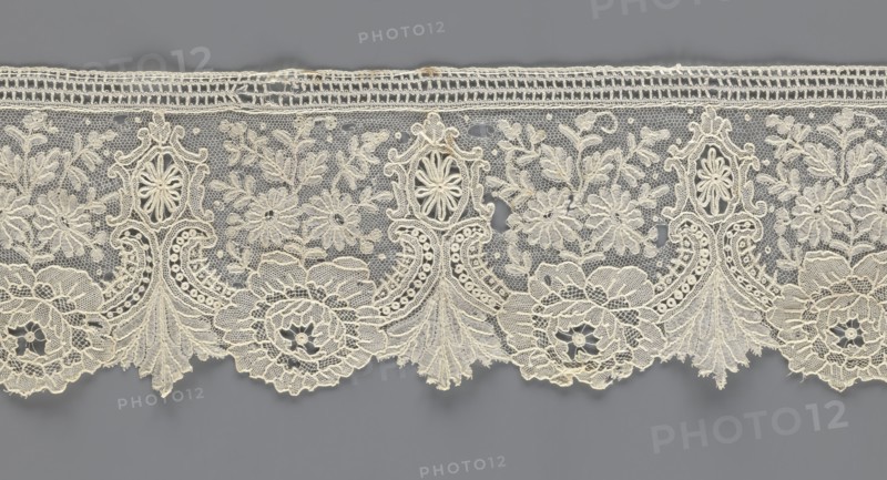 Lace fragment;, purchased provenance noted; lace with plant ornaments. Serrated on one side, with a pattern of flowers, fan-shaped leaves, and ovoid rosettes above them. Rose flowers between the leaves, and sprigs of other small flowers above them. Background fond de gaze. 2nd half of the 19th century, Brussels. Lace; width 8.5 cm, length 127 cm., A horizontal strip of delicate lace in off-white and cream tones stretches across a muted gray background, featuring an intricate repeating floral and ornamental pattern with scalloped lower edge; the lace shows a mesh-like netting filled with embroidered flowers, leaves, and ornamental medallions, with several larger floral rosettes and a few ornate vertical motifs that rise from the scallops, connected by swirling embroidered lines and tiny circular details, and the top edge has a narrow woven band in the same cream color.
