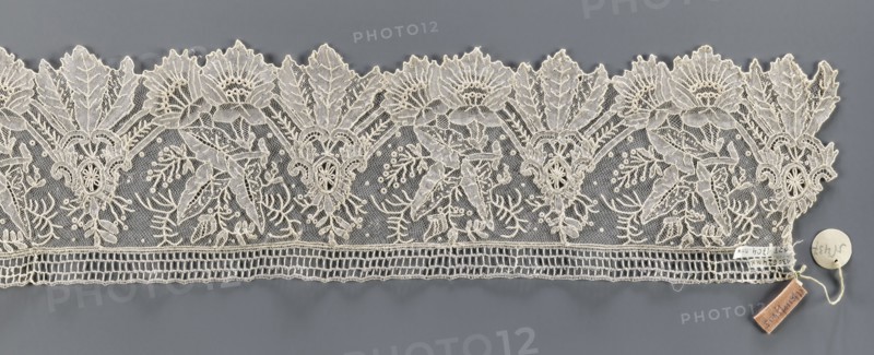 Lace fragment, Plant ornaments; pattern of stylized leaves and flowers, serrated on one side. Background fond de gaze. 2nd half of the 19th century, Brussels. Lace; width 11 cm, length 147 cm., A horizontal strip of lace with an intricate floral and leafy pattern laid flat on a gray background; the lace is off-white or cream in color and features repeating scalloped upper edges with large stylized flowers and pointed leaves, detailed openwork netting filling spaces between motifs, and denser embroidered outlines that create textured petals and foliage. Along the lower edge the lace has a band of rectangular openwork and a narrow embroidered border. A small round tag and a rectangular cloth tag attached by a thin string rest on the gray surface near one end of the lace; the visible colors are cream, gray, and a small area of muted brown on the rectangular tag.