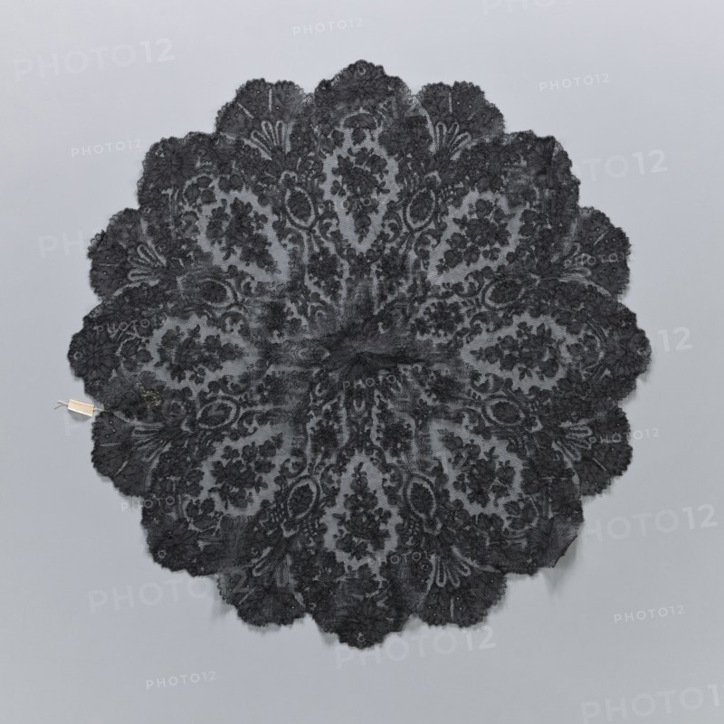 A fragment of lace umbrella cover, lace; patterns arranged radially from a central rosette; outer edge serrated with alternating fan-shaped leaves and large flower rosettes; above the rosette, leaves and C shapes frame an oval field containing a branch with flowers and rosebuds; 19th century, France; diameter 60 cm., A circular lace textile with a scalloped edge displayed flat on a light gray background, featuring an intricate floral and paisley-like pattern radiating from the center; the lace appears predominantly black with varying densities creating lighter gray areas where the background shows through, and there is a small pale rectangular tag attached near the outer edge.