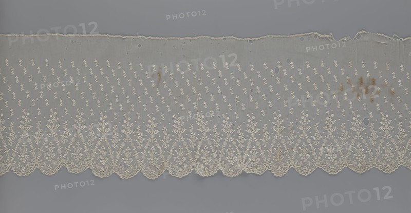 Lace fragment — shuttlecock; lace; clothing accessory; fashion. Lower edge: semicircular serrations bordered with fine serrations with geometric ornament. From the semicircular serrations upwards: a floral ornament repeating with a twig motif in triangular divisions; above this, a small twig in a regular projection. Mid-19th century, France. Material: lace. Width 42 cm, length 430 cm., A horizontal strip of delicate fabric with ornate embroidery lies against a flat background. The fabric is semi-transparent with a scalloped lower edge featuring dense floral and leafy embroidered motifs that form triangular arches along the border. Above the border, the textile has a repeating pattern of small floral sprigs scattered evenly across the field. The top edge is irregular and slightly frayed with several small holes visible throughout the upper portion of the fabric. There are scattered brownish stains on the right side and a few smaller discolorations elsewhere. The visible colors include o...