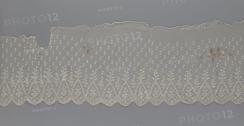 Lace fragment - shuttlecock, mid-19th century, France; lace; clothing accessory shuttlecock; lower edge with semicircular serrations bordered by fine serrations with geometric ornament; above semicircles a repeating floral ornament with a twig motif in triangular divisions; above this a small twig in a regular projection; width 42 cm, length 430 cm., A length of delicate lace fabric laid flat against a solid background, showing a scalloped lower edge with intricate floral embroidery and repeating ornate motifs above; the lace is predominantly off-white or cream with small embroidered leaf-like patterns across the body and denser floral designs along the scalloped border, and there are a few irregular holes and tears near the top edge as well as some small brownish stains scattered in several spots; the background behind the lace is a uniform cool gray.