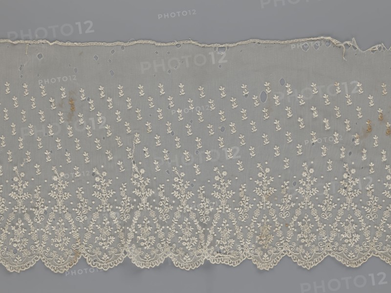 Lace fragment - shuttlecock;, purchase, Lace shuttlecocks used as clothing accessories and fashion. Lower edge: semicircular serrations bordered with fine serrations with geometric ornament. From the semicircular serrations upwards: floral ornament repeating with a twig motif in triangular divisions. Above this: a small twig in a regular projection. Mid-19th century, France. Material: lace. Width 42 cm, length 430 cm., A length of delicate lace fabric laid flat showing a sheer gray background with an overall pattern of small floral motifs embroidered in off-white or cream, a scalloped embroidered lower edge with denser, more intricate floral and vine designs, a worn upper edge with fraying, several small holes and irregular open areas scattered across the fabric, a few faint brownish stains, and subtle variations in thread texture and density.