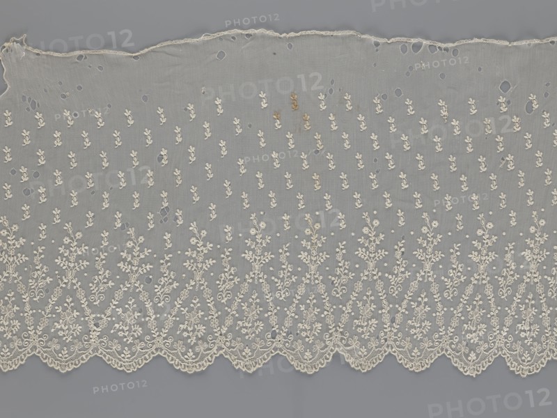 Lace fragment - shuttlecock, mid-19th century, France. Lace shuttlecock used as a clothing accessory/fashion item. Lower edge: semicircular serrations bordered with fine serrations with geometric ornament. From the semicircular serrations upwards: a floral ornament repeating with a twig motif in triangular divisions; above this, a small twig in a regular projection. Materials: lace. Dimensions: width 42 cm, length 430 cm., A length of delicate off-white lace fabric with a scalloped bottom edge and an irregular top edge, laid flat against a gray background; the lace has a repeating floral and leafy embroidered pattern that becomes denser toward the scalloped edge, with a sheer net-like ground showing a few small holes and some tiny darker spots; the visible colors are off-white for the lace and shades of gray for the background and the sheer areas.
