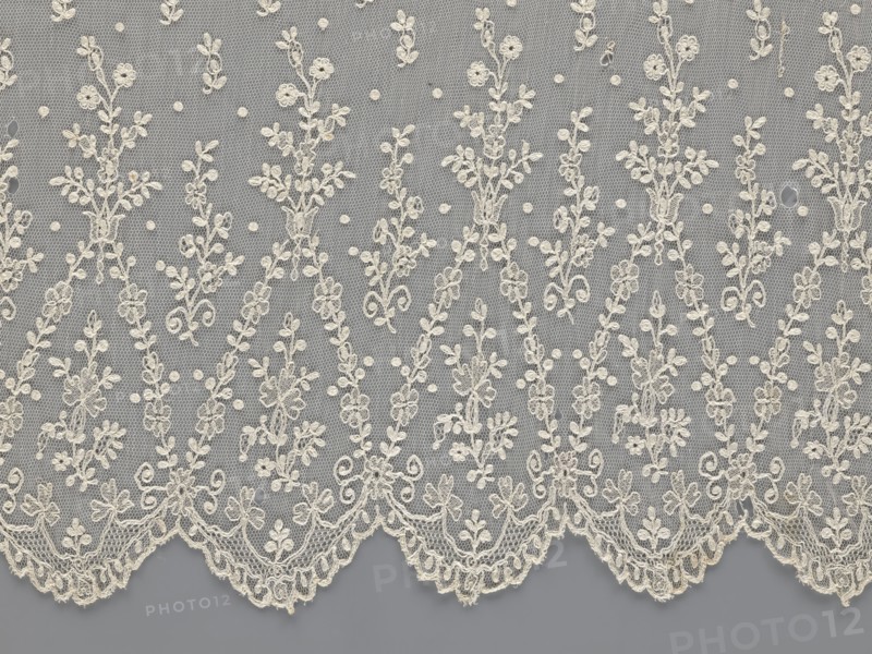 Lace fragment — shuttlecock; lace; clothing accessory; fashion. Lower edge: semicircular serrations bordered with fine serrations with geometric ornament. From the semicircular serrations upwards: a floral ornament repeating with a twig motif in triangular divisions. Above this, a small twig in a regular projection. Mid-19th century, France. Lace. Width 42 cm, length 430 cm., A delicate piece of lace fabric with an overall gray background and off-white or cream embroidered floral motifs, showing a repeating pattern of vertical floral sprays and small dot accents on a fine hexagonal mesh; the lower edge has a scalloped border with ornate scrollwork and floral clusters, and the embroidery includes leaves, tiny blossoms, and curling tendrils in the off-white or cream color against the gray.