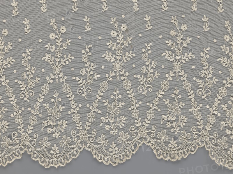 Lace fragment - shuttlecock;, mid-19th century, France. Lace shuttlecocks, clothing accessories, fashion. Lower edge: semicircular serrations bordered with fine serrations with geometric ornament. From the semicircular serrations upwards: a repeating floral ornament with a twig motif in triangular divisions; above this, a small twig in a regular projection. Materials: lace. Dimensions: width 42 cm, length 430 cm., A length of delicate lace fabric with an overall beige or off-white embroidered floral pattern set against a gray background, showing a repeating design of vertical vine-like motifs with small flowers and leaves connected by dotted netting, and a scalloped lower edge featuring denser floral embroidery and ornamental loops.