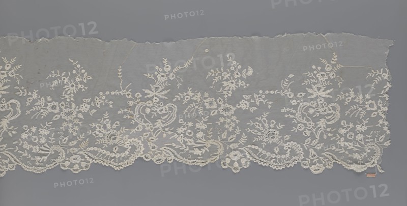 Lace fragment — shuttlecock; maker, purchase provenance; fashion, clothing accessory; needle lace and bobbin lace shuttlecocks; tiny spiders — needlework and bobbin ornament elements on factory-made tulle; floral ornament with a large repeat; serrated lower edge with stylized feathery leaves and flower calyces, above them floral bouquets and flower projections; 1st half of the 19th century, France; lace; width 39 cm, length 328 cm, A length of delicate lace fabric laid flat against a smooth background, showing an ornate floral and scrollwork embroidered border with scalloped lower edge; the lace is primarily off-white or cream with intricate motifs of flowers, leaves, vines, and looping paisley-like shapes, set on a fine translucent netting that appears pale gray where visible, and the background around the lace is a uniform medium gray.