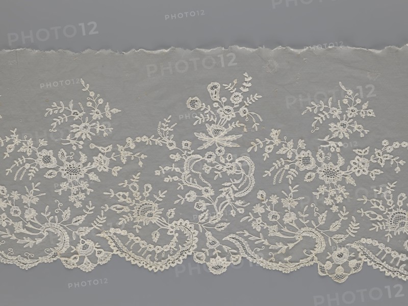 Lace fragment — shuttlecock; creator, purchase provenance; fashion; clothing accessories; lace; needle lace; bobbin lace; shuttlecocks; tiny spiders — needlework and bobbin ornament elements on factory-made tulle; floral ornament with a large repeat; serrated lower edge with stylized feathery leaves and flower calyces; above them floral bouquets and flower projections; 1st half of the 19th century; France; lace; width 39 cm; length 328 cm, A length of delicate lace fabric with an ornate floral and foliate pattern laid flat against a plain background; the lace shows a scalloped lower edge with curved motifs and a few openwork areas, and the upper edge appears slightly torn or uneven. The design features a group of embroidered flowers, leaves, tendrils, and small circular elements arranged in a repeating, asymmetrical layout, with sections of net-like mesh between the motifs. The visible colors are shades of off-white for the lace and a muted gray for the background.