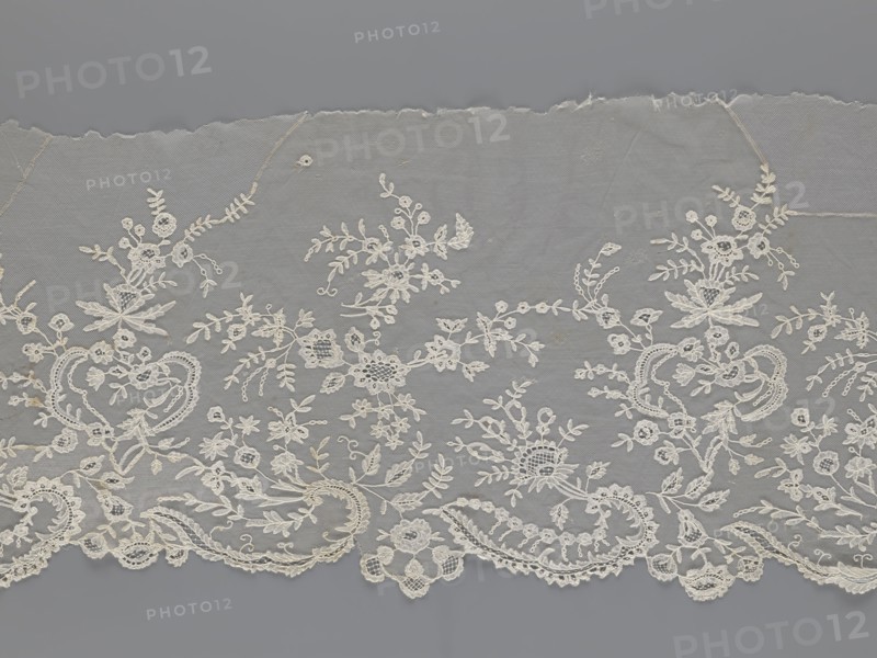 Lace fragment — shuttlecock;, Purchased provenance. Fashion accessory; needle lace and bobbin lace; shuttlecocks. Tiny spiders — needlework and bobbin ornament elements on factory-made tulle. Floral ornament with a large repeat. Serrated lower edge with stylized feathery leaves and flower calyces; above them are floral bouquets and flower projections. 1st half of the 19th century, France. Lace; width 39 cm, length 328 cm., A length of delicate lace fabric in shades of white and off-white lies against a gray background; the lace features an intricate floral and foliate pattern with a scalloped lower edge, showing clusters of stylized flowers, leaves, tendrils, and ornamental loops arranged in a repeating, flowing design, with some areas of slightly darker off-white thread and subtle shadowing that emphasize texture and raised embroidery.