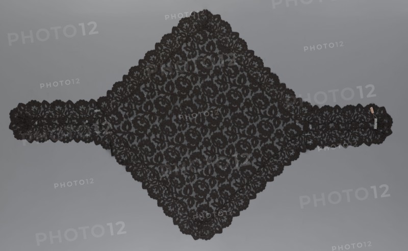 Cape, lace, in the form of a square with two wide, elongated lace ties. Continuous floral ornament with a motif of a large, asymmetrical flower sprig. Raised edge, a strip of floral edge ornament made separately and sewn on. 2nd half of the 19th century, France. Height 97 cm, width 168 cm., A large triangular piece of black lace with an intricate floral pattern lies flat against a neutral gray background, extending into a narrow band on each side; the lace has scalloped edges and repeating flower and leaf motifs, with the black lace appearing slightly raised against the gray, and a small neutral-colored tag attached near one end.