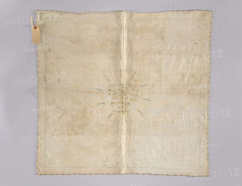Church napkin. Napkin made of linen with a fairly dense weave. In the middle an inscription embroidered with gold thread on a silk ribbon: Whoever eats my body / and drinks my blood has / eternal life And / I will raise / the last / day / in John in the year 6 / in 54. Around it is an appliquéd narrow grosgrain ribbon in cream and beige forming a circle with three-pronged rays 10, and within them are small blue and gold embroidered rays. An identical grosgrain ribbon in three tones of blue is sewn around the edges of the napkin. The ribbons, approximately 8 mm wide, date from the second half of the 18th century, but the inscription was created earlier, as the sewn-on ribbon partially obscures its lower letters. Fabric density: 30 warp and weft threads per cm. 18th century, Poland. Materials: linen, silk ribbon, gold thread, grosgrain. Dimensions: height 76 cm, width 80 cm., A square textile laid flat on a light gray background, appearing off-white to cream in color with areas of subtle...