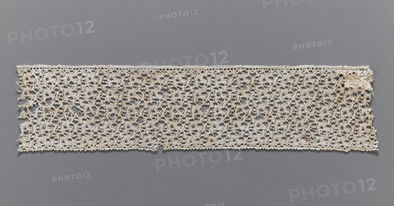 Lace fragment, bobbin lace, one edge finished with picots, 17th century, Flanders; width 6.5 cm, length 31 cm., No background. Repossession, A rectangular piece of openwork lace laid flat against a smooth background, showing an intricate repeating pattern of interconnected loops and small circular openings; the lace appears off-white to beige with subtle darker brownish spots or stains in places, and the background is a solid medium gray; edges of the lace are slightly irregular with a few frayed threads and tiny loose ends, and the lace surface shows slight variations in tone and texture across its length.
