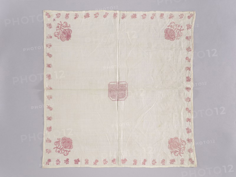 Velum, church paraments, Henryków German: Heinrichau. Flowers, leaves, plant ornaments; sacred art; churches and chapels furnishings. Velum made of white linen batiste. In the center, in a cartouche, an embroidered inscription in German and the date 1700. Border of small floral sprigs, with one large floral sprig in each corner. In one corner, a small monogram EN under a crown. Color of the ornament light raspberry. Flat, pierced, double-sided embroidery. Stitches: satin, cord, split, cross. Letters and numbers are made with crosses, each cross on three warp threads and three weft threads. 1700, Silesia. Liturgical vestment. Height 77 cm, width 75 cm., A square cloth laid flat on a light gray surface, mostly off-white in color with a visible central vertical and horizontal fold, featuring a delicate border of small floral motifs in pink along all edges and larger pink floral corner designs in three corners, with a pink decorative motif near the center; the fabric shows slight wrinkles ...