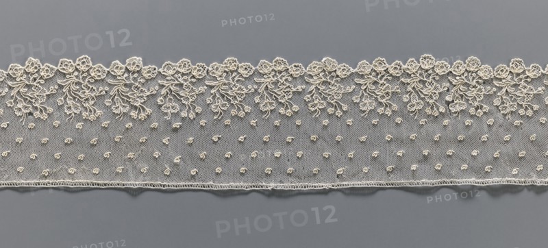 Lace fragment, repossession provenance; needle lace, Alençon lace, 18th century, France. The edge features a broad floral ornament composed of a repeated, elaborate bouquet of flowers; the edge is serrated following the forms of the flower calyxes, repeated in threes. Above, against the mesh background, very small heart-shaped leaves are depicted. Lace; width 8 cm, length 51 cm., A horizontal strip of delicate lace in off-white is laid against a smooth gray background; the lace features a scalloped top edge with an ornate floral and leafy embroidery pattern and a lower edge finished with a fine stitched border, while the central area of the lace is a fine net dotted with a few small raised embroidered buds.