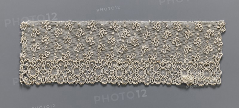 Lace fragment, needle lace, Alençon lace, one-sided geometric edge ornament with alternating ovals and quatrefoils; above, against a mesh background, asymmetrical twigs and dots are projected. 18th century, France. Lace; width 9 cm, length 29 cm., A rectangular piece of delicate lace displayed against a gray background, showing a fine hexagonal net ground in a beige or off-white color with a scalloped lower edge, an ornate lower border of interconnected oval and floral loop motifs in beige with raised corded outlines, and a field above scattered with a few small looped floral sprigs and dots in the same beige tone; the overall palette is beige and off-white set on gray.