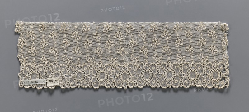 Lace fragment, needle lace, Alençon lace, one-sided geometric edge ornament including alternating ovals and quatrefoils; above, against a mesh background, asymmetrical twigs and dots are projected. 18th century, France. Width 9 cm, length 29 cm., Repossession, A rectangular piece of delicate lace displayed flat against a gray background, showing a fine hexagonal net ground with a pattern of small clustered floral motifs and raised dots across the field; the lower edge features a decorative scalloped border of interconnected oval and circular motifs with complex looping outlines and openwork, while the top edge is straighter and more irregular; the lace is an off-white or creamy color contrasting with the medium gray background.