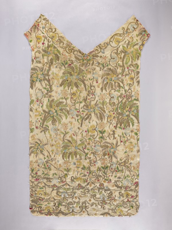 Back of the chasuble. Back of the chasuble made of cream silk rep, embroidered with patterns of various flower branches, palm trees, leaves, no specific composition. Probably adapted into a chasuble from another object. Embroidery with colored silk thread using flat, satin stitches and smooth and slightly crimped gold thread. Gold and pastel colors: blue, green, yellow, red, pink. 17th/18th century, Poland. Liturgical vestment. Length 133.5 cm, width 72 cm., A sleeveless textile garment with a V-shaped neckline displayed flat on a pale background, covered in an all-over floral and foliate woven pattern; the fabric is primarily cream and beige with visible accents of green, yellow, blue, pink, and touches of brown, showing stylized flowers, leafy fronds, and curling vine motifs embroidered or woven throughout, with a scalloped decorative border along the lower edge and a symmetrical layout that tapers toward the shoulders.