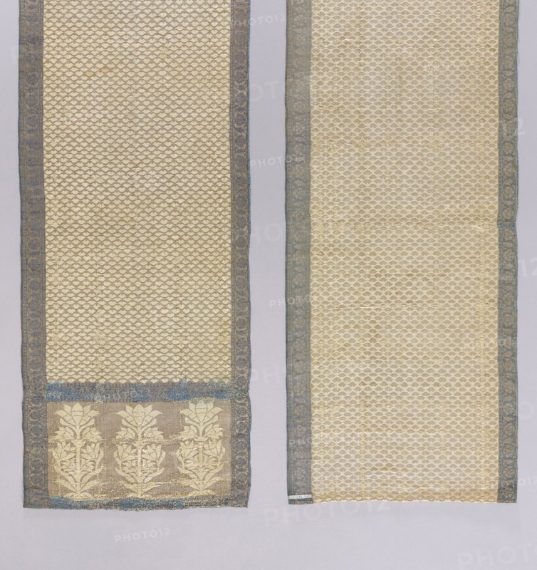 Kontusz belt, Tulips; double-sided, semi-solid triple-section heads with stylized tulip flowers and alternating boat motifs; floral tendril border; double triangle borders; geometric and plant ornaments. Right side: silver background, white ornament on heads and central part, navy blue borders; left side is the reverse. Unsigned; type associated with Istanbul/Constantinople belts—edge weaving and double-triangle motif characteristic of Armenian belts; making method similar to Constantinople examples; similar to a French or Gdodno belt with the same bush-and-flower motif. First half of the 18th century. Poland culture. National costume accessory. Length 268 cm; width 32 cm., A pair of long rectangular textile panels displayed side by side on a pale background, both panels showing an overall cream-colored ground with a repeating small-scale scalloped or scale-like pattern and narrow blue-gray decorative borders along their long edges, the left panel having a wider lower section with a ba...