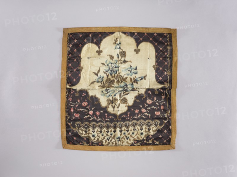 Tablecloth; maker, church paraments; printed fabric with flowers, leaves, and plant ornaments; sacred art for churches and chapels furnishings; 19th century; material: fabric; height 63.5 cm, width 59.8 cm, A square fabric panel with a golden brown border lies flat against a light gray background, showing a central cream-colored cartouche containing a floral motif in muted blue and brown tones, surrounded by a dark brown field patterned with small pink floral elements and lattice-like accents, with additional cream and brown decorative bands and a scalloped border design near the bottom.