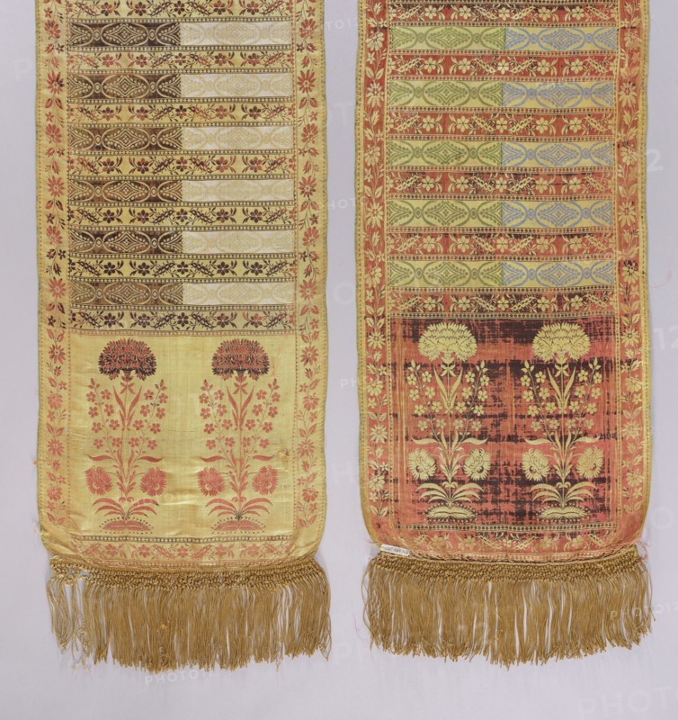 Kontusz belt, Bipartite heads with a motif of elongated flower bushes of various species with carnations at the top, alternating transverse stripes with a plant twig and a motif of elongated rosettes with rhombuses. Floral twig border. Sewn-in silk fringe, yellow, braided at the top. Colors: straw, brown, white, green, and blue. At the bottom of one of the heads a lying FS preserved. 2nd half of the 18th century, Kraków, national costume, clothing accessory. Length 396 cm, width 38 cm., Two long narrow woven textile panels hang side by side, each with a decorative border and fringe at the bottom. The left panel has a pale yellow ground with horizontal patterned bands near the top in darker tones and a floral motif in coral and brown below, framed by a coral floral border and finished with a gold fringe. The right panel has a warmer, more reddish background with greenish and bluish horizontal patterned bands and a darker rectangular area near the lower portion featuring cream floral mot...