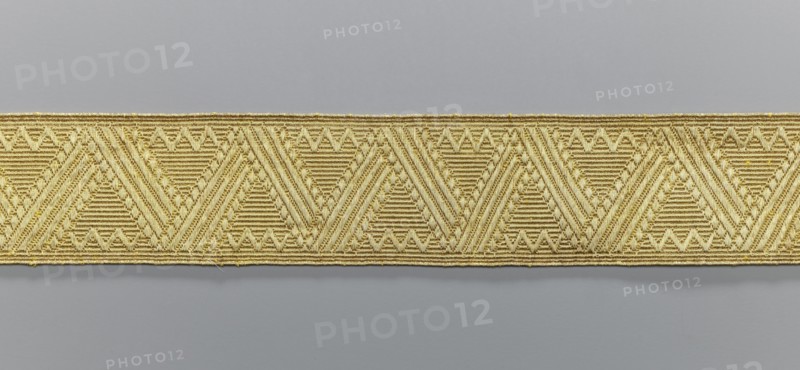 Gallon, origin unknown, galloons haberdashery, geometric ornaments, smaller snake pattern at the edges, early 19th century, Europe, fabrics, width 4.7 cm, length 53 cm, A horizontal strip of woven trim with a repeating geometric pattern sits against a smooth background; the trim shows a series of triangular motifs formed by tightly woven ridged lines and small raised accents, creating a textured surface with diagonal and horizontal linear detailing, and the visible colors are gold and gray.