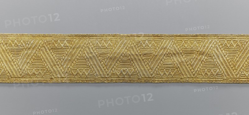 Gallon, galloons haberdashery, provenance unknown, geometric ornaments, a smaller snake pattern at the edges, early 19th century, Europe, fabrics, width 4.9 cm, length 52 cm, A horizontal woven trim with a repeating geometric pattern runs across the image, showing a group of chevron and triangular motifs formed by parallel textured lines; the trim appears gold with variations of lighter and darker gold tones against a neutral gray background.