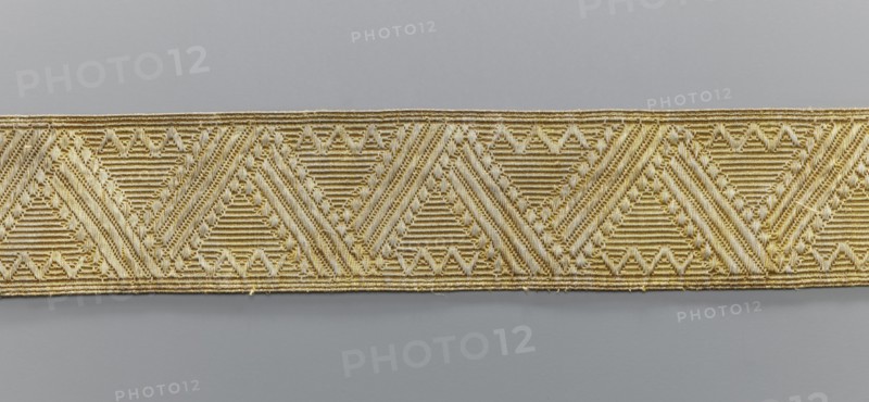 Gallon, galloons haberdashery, origin unknown, geometric ornaments, smaller snake pattern at the edges, early 19th century, Europe, fabrics, width 5.1 cm, length 41.8 cm, A narrow woven band with a repeating geometric pattern showing a row of connected triangular motifs formed by alternating diagonal and horizontal ribbed textures; the band appears in shades of gold and beige against a smooth light gray background.