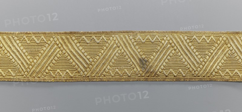 Gallon, galloons haberdashery, origin unknown, geometric ornaments, smaller snake pattern at the edges, early 19th century, Europe, fabrics, width 5.1 cm, length 41.8 cm, A narrow decorative band runs horizontally across the image, featuring a repeating geometric pattern of triangular motifs and diagonal ribbed textures. The band is primarily gold in color with variations of lighter and darker golden tones, and it has a slightly shiny, metallic sheen. The background above and below the band is a smooth, muted gray with subtle tonal variation. The gold band shows some areas with faint darker discoloration and small irregularities in the threads, and the edges of the band are slightly uneven with tiny loose fibers.