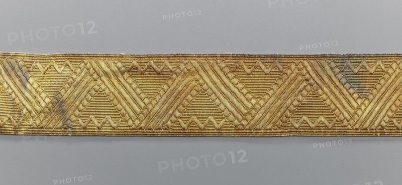 Gallon, galloons haberdashery, origin unknown, geometric ornaments, smaller snake pattern at the edges, early 19th century, Europe, fabrics, width 5 cm, length 53.6 cm, A horizontal strip of woven fabric with a repeating geometric pattern dominated by triangular and diagonal motifs in metallic gold tones sits against a neutral gray background; the fabric shows variations of bright gold and darker antique gold with small areas of muted grayish discoloration, and the woven texture reveals parallel ridges and raised braided details throughout the strip.